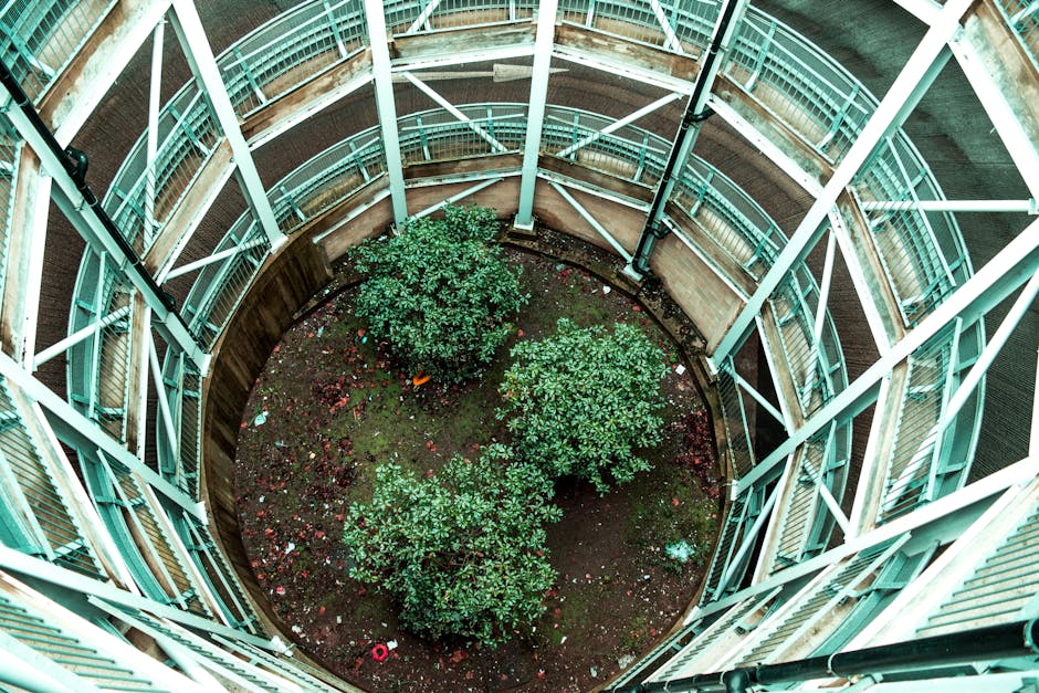 A circular garden courtyard surrounded by multi-storey residential buildings with external walkways and metal railings painted in light turquoise. The garden features a central patch of dark, moist so