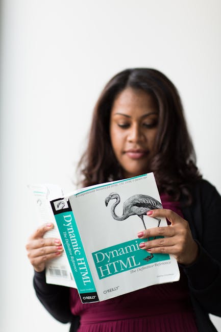 The image depicts a woman with shoulder-length dark hair, dressed in a dark blazer over a maroon top, standing indoors against a plain light background. She is focused on reading a book titled 'Dynami